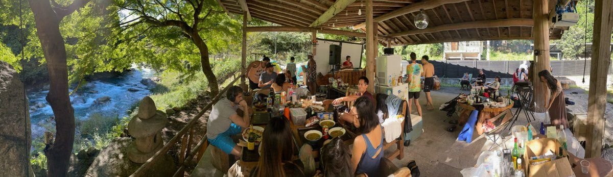 A group of people gather under a wooden pavilion by the river, reminiscent of a scene at Nikko Hostel, enjoying a meal at tables filled with food and drinks. Trees and the serene river are visible in the background.